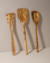 A set of three olive wood kitchen utensils, including a flipper, a pointed spoon, and a pancake spatula, laid out on a neutral surface.